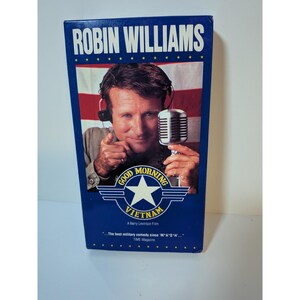 Peut inclure: Une cassette VHS rectangulaire avec une bordure bleue et une image centrale de Robin Williams portant un casque et tenant un microphone. Le texte indique ROBIN WILLIAMS, GOOD MORNING VIETNAM, A Barry Levinson Film, et The best military comedy since M*A*S*H.