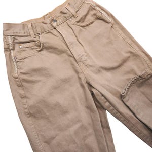 May include: High-waisted tan denim jeans featuring a button and zip fly closure, belt loops, and five pockets. The front pockets and side seams are accented with decorative white cross-stitch embroidery and silver-toned metal rivets.
