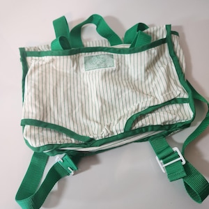 May include: A white backpack with thin green vertical stripes, featuring green nylon webbing straps, green trim along the edges, and a rectangular green fabric label centered on the front. The bag includes adjustable straps with white plastic buckles.