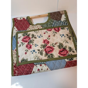 Puede incluir: Un bolso de tela acolchada con un diseño de patchwork de motivos florales en rojo, rosa y azul sobre un fondo crema. El bolso cuenta con un bolsillo exterior rectangular y dos asas de madera clara integradas en el borde superior.