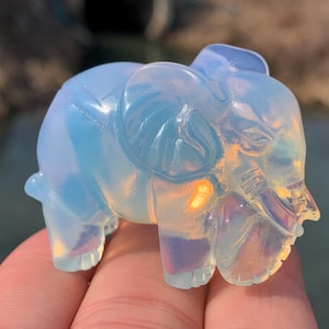 Pode incluir: Uma pequena figura de elefante de opala branca com um acabamento translúcido e iridescente. A figura é esculpida com detalhes precisos, incluindo a tromba, as orelhas e os olhos do elefante.