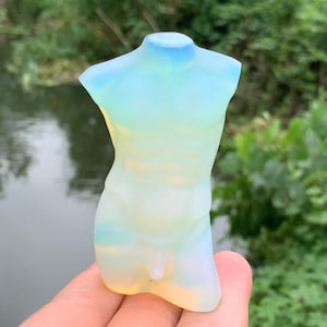 Peut inclure: Une sculpture de torse translucide et opalescente avec un dégradé de couleurs. La sculpture passe d'un bleu clair au niveau du cou à un jaune doux à la base. La forme est tenue dans une main sur fond de feuillage vert et d'eau.