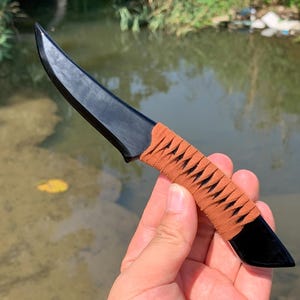 Può includere: Un coltello nero con una lama curva e un manico avvolto in marrone. Il coltello è tenuto contro uno sfondo d'acqua, mostrando il suo design elegante e la sua maestria. La lama appare affilata e pronta all'uso.