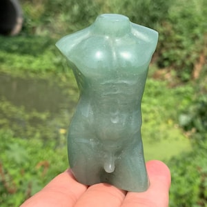 Peut inclure: Une sculpture de torse en pierre précieuse verte sculptée. La figurine est une représentation stylisée de la forme masculine, avec une musculature définie. La sculpture est tenue dans une main sur un fond vert flou.