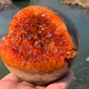 May include: A wooden sphere with a large, orange crystal cluster embedded in one side. The sphere is dark brown on the outside and has a natural wood grain pattern.