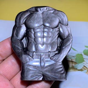 Peut inclure: Une figurine de torse sculpté grise avec des muscles définis, notamment la poitrine, l'abdomen et les bras. La figurine porte un pantalon avec une ceinture. La figurine est tenue dans une main sur un fond jaune et un livre.