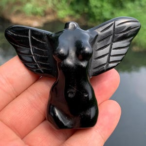 Peut inclure: Une figurine d'ange en obsidienne noire avec des ailes sculptées. La sculpture représente un torse féminin avec des ailes déployées. Les ailes présentent des sculptures de plumes détaillées. La figurine est tenue dans une main sur un fond flou.