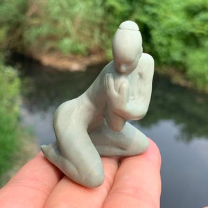 Peut inclure: Une figurine vert clair représentant une personne agenouillée, les mains jointes devant la poitrine. La figurine a une coiffure en chignon et est agenouillée sur les deux genoux. La figurine est tenue dans une main, avec un arrière-plan flou de feuillage vert et d'eau.
