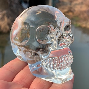 May include: A clear glass skull sculpture with detailed features, including eye sockets, a nose, and teeth. The skull is sitting on a hand.
