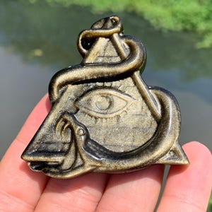 Puede incluir: Colgante de obsidiana dorada con una pirámide con un ojo y una serpiente. El colgante tiene un diseño detallado y un acabado pulido. El fondo está borroso, lo que sugiere un entorno al aire libre.
