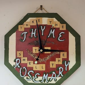 May include: A red, green, and white octagonal clock with a black clock face and hands. The clock face is surrounded by wooden letter tiles spelling out "Take the Thyme to Smell the Rosemary".