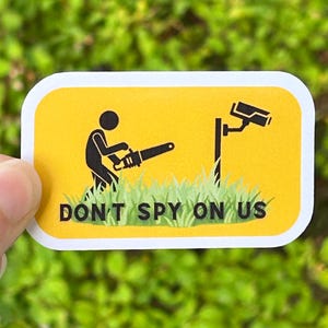 May include: A rectangular sticker with a yellow background featuring a black stick figure holding a chainsaw toward a security camera mounted on a pole. Green grass is depicted at the bottom with the black text DON'T SPY ON US printed below.
