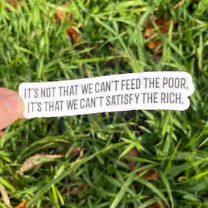 May include: A white sticker with black text: "IT'S NOT THAT WE CAN'T FEED THE POOR, IT'S THAT WE CAN'T SATISFY THE RICH." The sticker is held against a background of green grass.