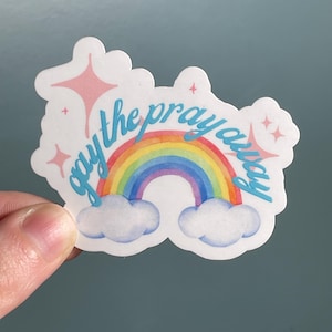 May include: A sticker with a rainbow design and the words "gay the pray away" in light blue script. The sticker also features two white clouds and pink star accents.