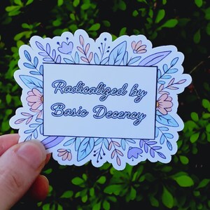 Puede incluir: Una pegatina con el texto "Radicalized by Basic Decency" en fuente cursiva, dentro de un rectángulo blanco. El rectángulo está rodeado por un borde floral en tonos pastel. La pegatina se sostiene sobre un fondo de follaje verde.