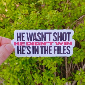 May include: A white sticker with black text that reads "HE WASN'T SHOT HE DIDN'T WIN HE'S IN THE FILES." The text "HE DIDN'T WIN" is in pink. The sticker is held in front of a background of green foliage.