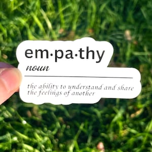 Könnte beinhalten: Ein weißer Aufkleber mit dem Wort "empathy" in Schwarz und der Definition "die Fähigkeit, die Gefühle anderer zu verstehen und zu teilen." Der Aufkleber befindet sich vor einem Hintergrund aus grünem Gras.