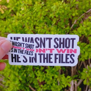 May include: Three white stickers with black text and red accents. The stickers read "HE WASN'T SHOT", "DIDN'T WIN", and "HE'S IN THE FILES". The background is blurred green foliage.