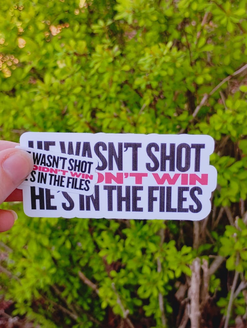 May include: Three white stickers with black text and red accents. The stickers read "HE WASN'T SHOT", "DIDN'T WIN", and "HE'S IN THE FILES". The background is blurred green foliage.