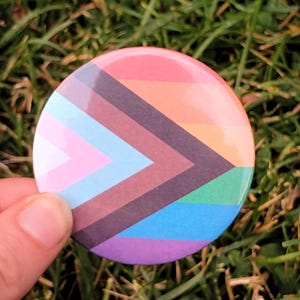 May include: A circular pin-back button featuring the Progress Pride flag design. The button displays horizontal stripes of red, orange, yellow, green, blue, and violet, overlaid with a chevron pattern in white, pink, light blue, brown, and black.