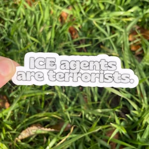 May include: A white sticker with black text that reads "ICE agents are terrorists." The sticker is held against a background of green grass.