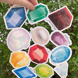 May include: A collection of gemstone stickers in various shapes and colors, including blue, green, red, and yellow. Each sticker has a month name written on it, such as "september" and "may". The stickers are held against a backdrop of green foliage.