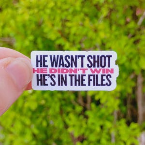 May include: A rectangular sticker with the text "HE WASN'T SHOT, HE DIDN'T WIN, HE'S IN THE FILES" in black and pink lettering. The sticker is held against a blurred green background.