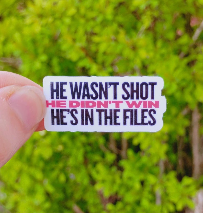 May include: A rectangular sticker with the text "HE WASN'T SHOT, HE DIDN'T WIN, HE'S IN THE FILES" in black and pink lettering. The sticker is held against a blurred green background.