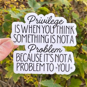May include: A white sticker with black text that reads "Privilege is when you think something is not a problem because it's not a problem to you." The sticker is held against a backdrop of green and yellow foliage.
