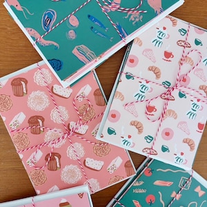May include: Four stacks of rectangular greeting cards tied with red and white baker's twine. The cards feature colorful patterns including sea life on green, pastries on pink, breakfast items on light pink, and pasta shapes on green backgrounds.