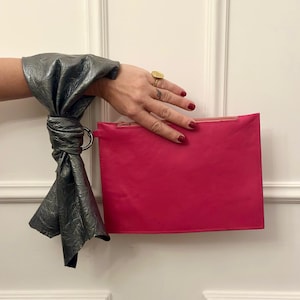 May include: A bright pink leather clutch with a zipper closure. The clutch is held by a hand wearing a silver leather bracelet with a ring.