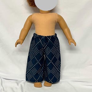 May include: A doll wearing a tan sleeveless top and navy blue wide-leg pants. The pants feature a white diamond pattern. The doll has red hair and is posed against a white background.