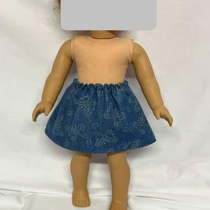 May include: A doll wearing a peach-colored sleeveless top and a blue denim skirt. The skirt features a pattern of white floral designs. The doll's skin tone is tan.