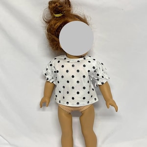 May include: A doll wearing a white blouse with black polka dots. The blouse has short, ruffled sleeves. The doll has red hair styled in a bun and is wearing beige underwear. The doll is standing on a white surface.