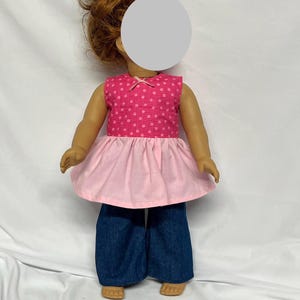 May include: A doll wearing a pink polka dot top with a light pink skirt and dark blue denim pants. The top has a small bow at the neckline. The doll's hair is styled in a bun.