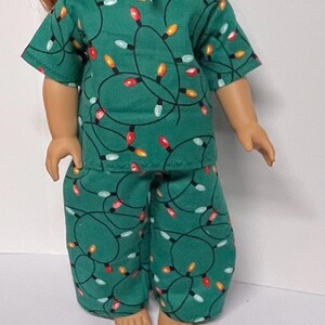 May include: Green doll pajamas set with a repeating pattern of colorful Christmas lights. The short-sleeved top and pants are made of a soft fabric. A white bow is at the top of the doll's head.
