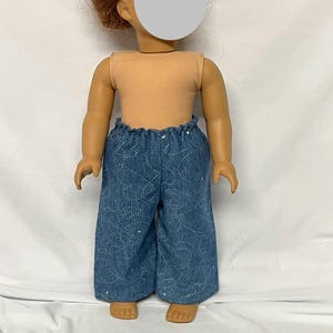May include: A doll wearing a tan sleeveless top and blue denim-style pants. The pants have a wide leg and are decorated with white stitching. The doll has red hair and is standing on a white surface.
