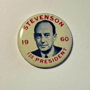 May include: A vintage campaign badge featuring a portrait of a man. The badge has the text "STEVENSON" in a circular pattern, with "1960" and "FOR PRESIDENT" also printed on it. The badge is off-white with red text.