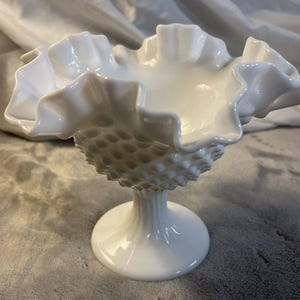 Puede incluir: Un cuenco de vidrio blanco con borde ondulado y detalles de hobnail en la parte inferior. El cuenco se apoya en una base de pedestal. El diseño evoca un ambiente vintage, similar a la cristalería antigua. Es un objeto decorativo.