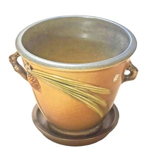 Puede incluir: Una maceta de cerámica con plato a juego, que presenta un esmalte en tonos ámbar y un diseño en relieve de agujas de pino y una piña. La maceta tiene dos asas laterales con forma de ramas y un borde interior liso de tono gris.