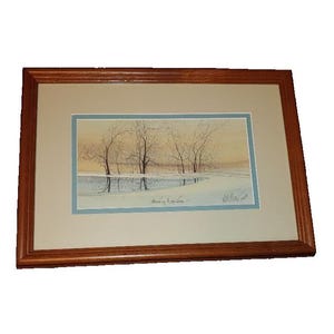 Könnte beinhalten: Gerahmtes Aquarell-Landschaftsbild mit kahlen Bäumen, die sich in Wasser unter einem sanften, goldenen Himmel spiegeln. Das Kunstwerk ist in einem hellblauen Passepartout und einem dunklen Holzrahmen gefasst, was eine ruhige Szene schafft.