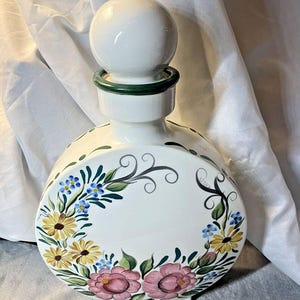 Puede incluir: Una garrafa redonda de cerámica blanca con un tapón esférico y un borde verde oscuro. El cuerpo presenta motivos florales pintados a mano, incluyendo margaritas amarillas, nomeolvides azules y flores rosas con hojas verdes y zarcillos negros.