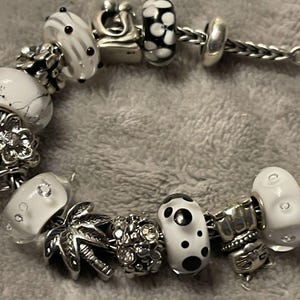 Puede incluir: Una pulsera de plata con varios abalorios decorativos. Las cuentas incluyen diseños blancos y negros, detalles florales y un charm de palmera. La pulsera está sobre una superficie gris.
