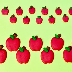 May include: A collection of red apple-shaped candies with green leaves and brown stems, arranged on a light green background. The candies vary in size, creating a playful and visually appealing pattern. The image has a bright and cheerful aesthetic.