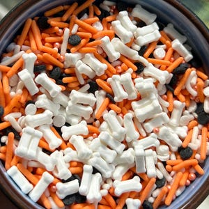 May include: A close-up shot of a bowl filled with Halloween-themed sprinkles. The mix includes white bone-shaped sprinkles, orange jimmies, black nonpareils, and small white and orange beads. The bowl has a blue and brown rim.