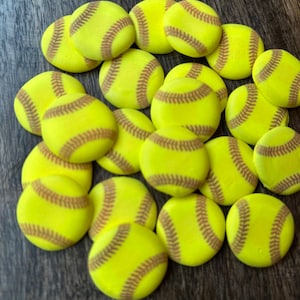 May include: A collection of small, yellow softball-shaped objects with brown stitching. The objects are arranged on a dark wooden surface. The softballs are a bright yellow color with detailed brown stitching, resembling real softballs.