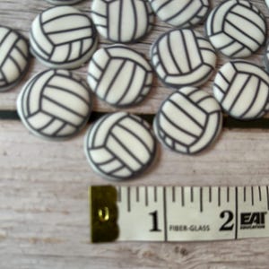 May include: A close-up of a group of white and black volleyball-themed cake decorations, each measuring approximately 1 inch in diameter. The decorations are arranged on a wooden surface with a measuring tape.