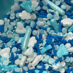 May include: A close-up of a mix of blue, white, and silver sprinkles. The sprinkles include small round shapes, long rods, and cloud and baby foot shapes. The color palette is a mix of light and dark blues, white, and silver.