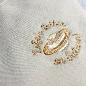 May include: White t-shirt with gold embroidery that reads "Life's better on Saturn" and features a stylized image of the planet Saturn.