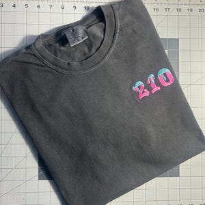 May include: A dark gray t-shirt with a pink and blue embroidered design that says "210".